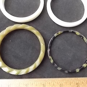 May include: Four bangles in the image. Two are solid white, one is a light green with a twisted design, and the last is black and green with a speckled pattern. The bangles are displayed on a dark gray surface.
