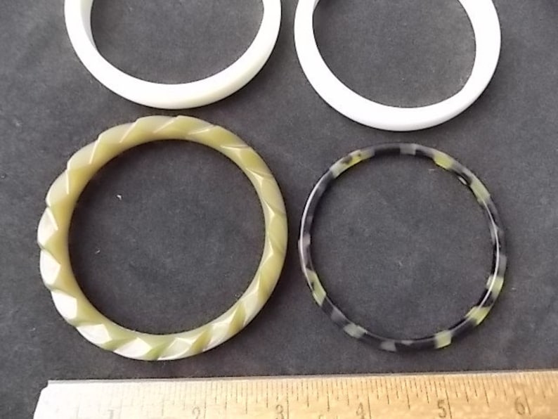 May include: Four bangles in the image. Two are solid white, one is a light green with a twisted design, and the last is black and green with a speckled pattern. The bangles are displayed on a dark gray surface.