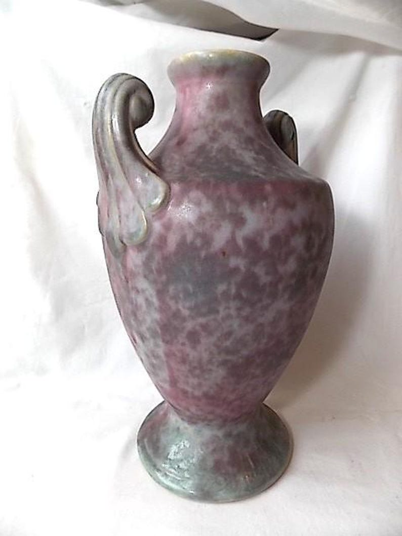 May include: A speckled, antique-style vase with a wide base and two curved handles. The vase has a pink and gray mottled pattern, with a light green rim and base. The overall shape is reminiscent of a classic urn.