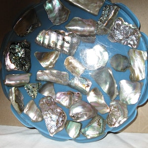 May include: A blue, scalloped-edge bowl decorated with iridescent abalone shells. The shells vary in size and shape, showing silver, green, and purple hues. The bowl's design creates a decorative and natural aesthetic.