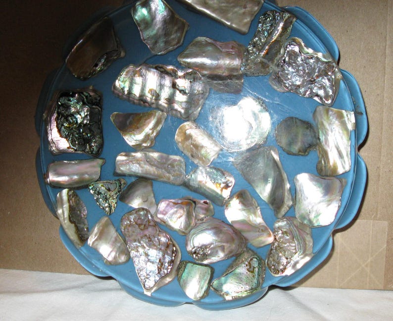 May include: A blue, scalloped-edge bowl decorated with iridescent abalone shells. The shells vary in size and shape, showing silver, green, and purple hues. The bowl's design creates a decorative and natural aesthetic.