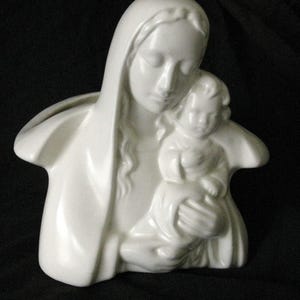 May include: A white ceramic sculpture of Mary holding the baby Jesus. The figures are depicted with serene expressions and flowing robes. The sculpture is a religious item, likely for home decor or display.