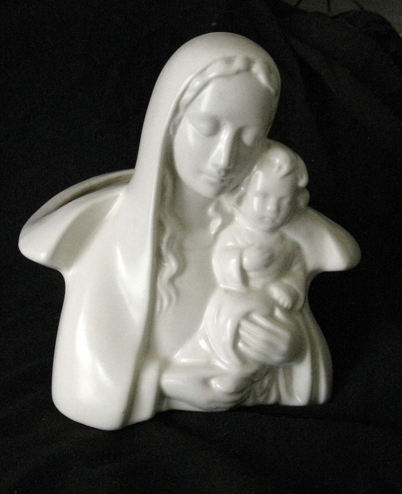 May include: A white ceramic sculpture of Mary holding the baby Jesus. The figures are depicted with serene expressions and flowing robes. The sculpture is a religious item, likely for home decor or display.