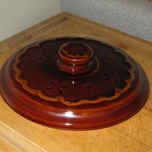 May include: A brown ceramic lid with a scalloped edge and a central knob. The lid features a floral design and is glazed with a glossy finish. The lid is likely for a casserole dish or similar kitchenware.