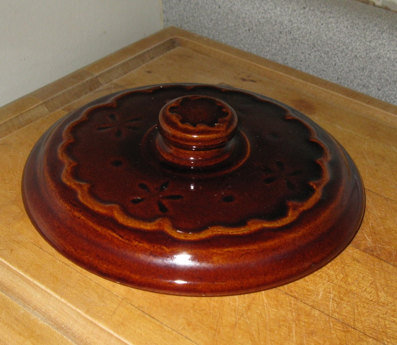 May include: A brown ceramic lid with a scalloped edge and a central knob. The lid features a floral design and is glazed with a glossy finish. The lid is likely for a casserole dish or similar kitchenware.