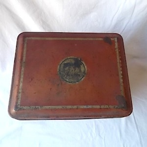 May include: A rectangular metal storage tin with a weathered, reddish-orange finish. The lid features a central circular emblem with a dark, indistinct graphic and a thin, faded gold-colored border framing the top surface of the container.