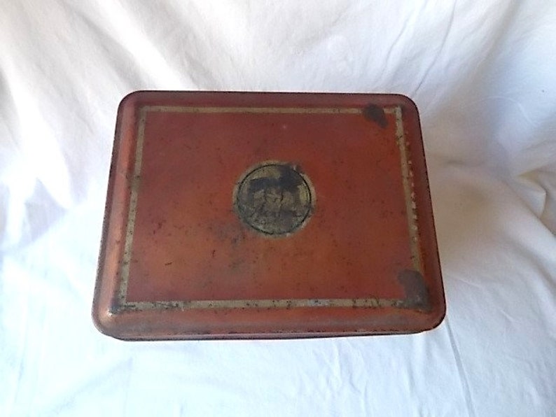 May include: A rectangular metal storage tin with a weathered, reddish-orange finish. The lid features a central circular emblem with a dark, indistinct graphic and a thin, faded gold-colored border framing the top surface of the container.