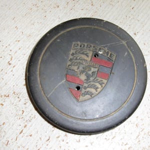 Puede incluir: Un emblema Porsche gris oscuro con el logotipo de Porsche y un dise&ntilde;o de escudo. El emblema tiene detalles en rojo y negro y dos peque&ntilde;os agujeros. El emblema circular parece ser de metal.