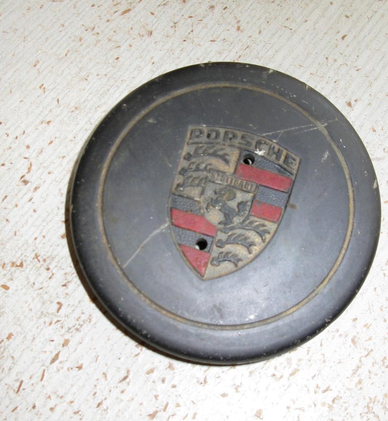 Puede incluir: Un emblema Porsche gris oscuro con el logotipo de Porsche y un dise&ntilde;o de escudo. El emblema tiene detalles en rojo y negro y dos peque&ntilde;os agujeros. El emblema circular parece ser de metal.