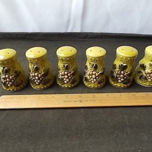 May include: A set of six vintage ceramic spice shakers. Each shaker is a light green color with a yellow top and features a grape design. The shakers are labeled with the names of different spices. A wooden ruler is placed in front of the shakers.