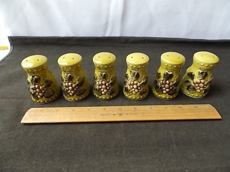 May include: A set of six vintage ceramic spice shakers. Each shaker is a light green color with a yellow top and features a grape design. The shakers are labeled with the names of different spices. A wooden ruler is placed in front of the shakers.