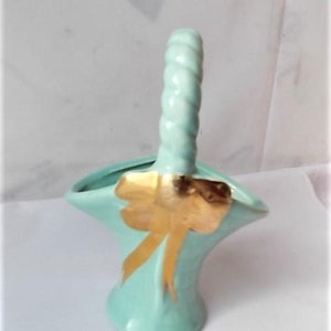 May include: A light green ceramic basket with a twisted handle and a gold bow design. The basket has a flared shape and is likely vintage. The gold bow adds a decorative touch to the basket.