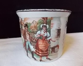 May include: A white ceramic planter featuring a colorful illustration of a person in a brown coat and top hat standing near a person in a white blouse, green vest, and red skirt. The rim of the planter has a dark, weathered metallic finish.