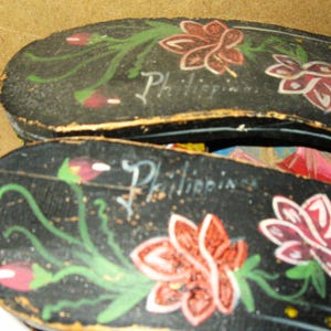 Peut inclure: Une paire de sandales en bois noir orn&eacute;es de motifs floraux peints &agrave; la main en rouge, blanc et vert. Le mot Philippines est inscrit en &eacute;criture blanche sur la surface de chaque sandale. La chaussure a une forme plate et allong&eacute;e.