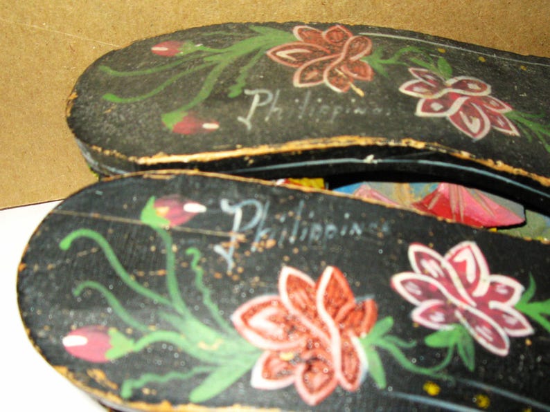 Peut inclure: Une paire de sandales en bois noir orn&eacute;es de motifs floraux peints &agrave; la main en rouge, blanc et vert. Le mot Philippines est inscrit en &eacute;criture blanche sur la surface de chaque sandale. La chaussure a une forme plate et allong&eacute;e.