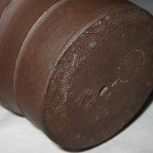 Peut inclure: Un r&eacute;cipient cylindrique en c&eacute;ramique brun fonc&eacute; avec une base plate. Le fond pr&eacute;sente une marque circulaire estamp&eacute;e avec du texte et un petit trou central. La surface ext&eacute;rieure a une texture mate et terreuse avec des stries horizontales.