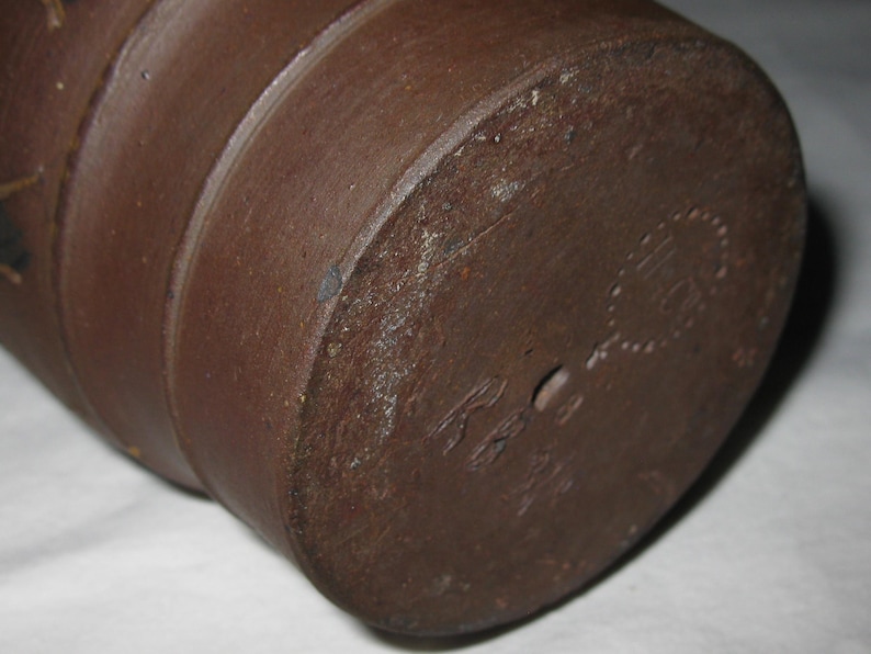 Peut inclure: Un r&eacute;cipient cylindrique en c&eacute;ramique brun fonc&eacute; avec une base plate. Le fond pr&eacute;sente une marque circulaire estamp&eacute;e avec du texte et un petit trou central. La surface ext&eacute;rieure a une texture mate et terreuse avec des stries horizontales.