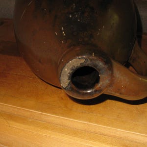 Pode incluir: Um jarro de cer&acirc;mica castanho com bico e asa. O jarro tem um acabamento brilhante e parece ser vintage. O bico &eacute; circular e a asa curva-se para cima. O jarro est&aacute; sobre uma superf&iacute;cie de madeira.