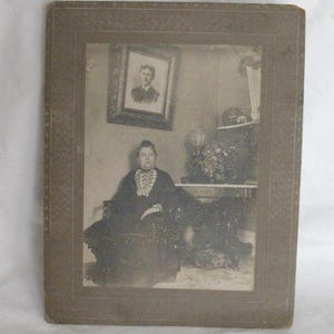 May include: Antique black and white photograph of a woman seated in a chair, wearing a dark dress with a lace collar. A framed portrait hangs on the wall behind her. The photograph is in a decorative frame.