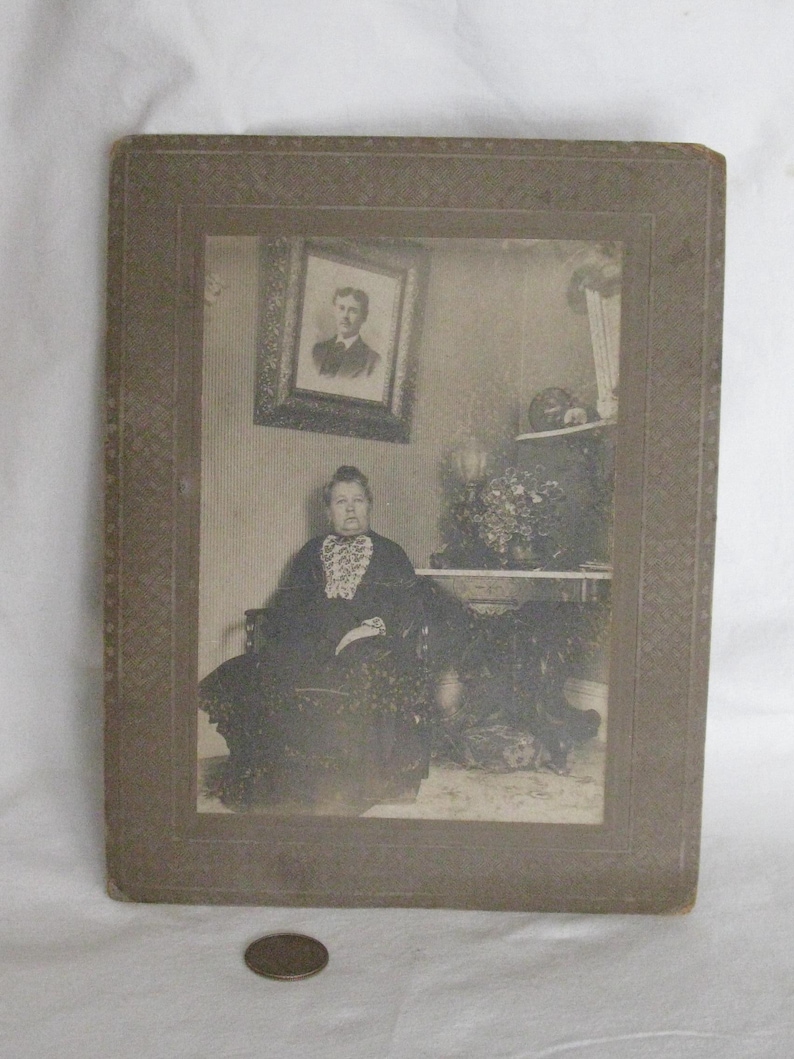 May include: Antique black and white photograph of a woman seated in a chair, wearing a dark dress with a lace collar. A framed portrait hangs on the wall behind her. The photograph is in a decorative frame.