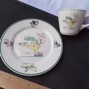 May include: A vintage children's dish set featuring a plate and a cup. The plate and cup are white with a green trim and illustrations of a cartoon rabbit ice skating. The plate has the name "DONALD" on it. The cup has the name "OSWALD" on it.