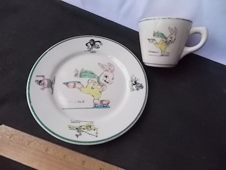 May include: A vintage children's dish set featuring a plate and a cup. The plate and cup are white with a green trim and illustrations of a cartoon rabbit ice skating. The plate has the name "DONALD" on it. The cup has the name "OSWALD" on it.
