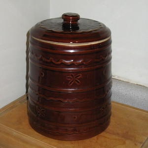 May include: A brown ceramic canister with a lid. The canister has a cylindrical shape with decorative horizontal bands and floral patterns. The lid has a knob and a scalloped edge. The overall design is rustic and vintage.