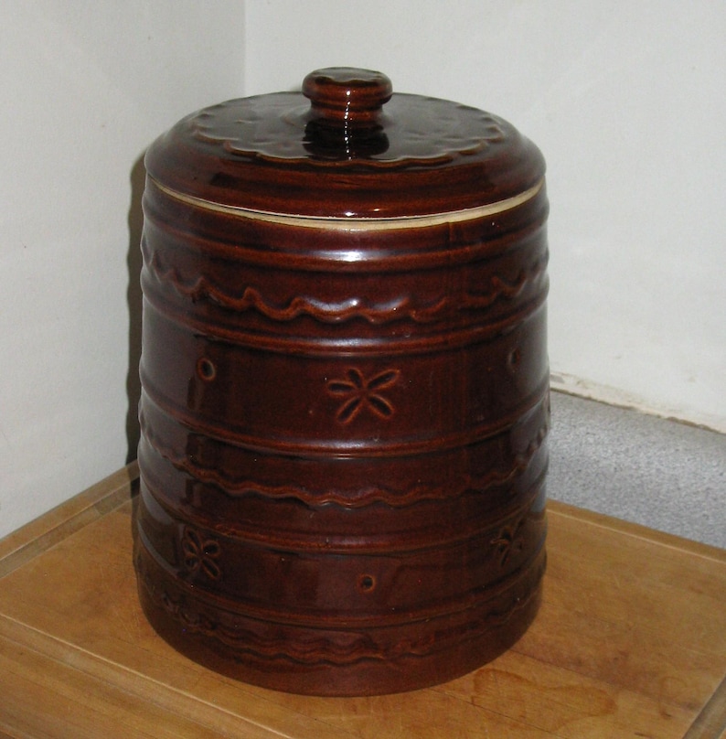 May include: A brown ceramic canister with a lid. The canister has a cylindrical shape with decorative horizontal bands and floral patterns. The lid has a knob and a scalloped edge. The overall design is rustic and vintage.
