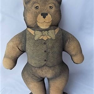 May include: A stuffed teddy bear with a light brown face, paws, and feet, and a darker brown body. The bear wears a vest with a herringbone pattern and a bow tie. The bear has button eyes and a stitched nose.