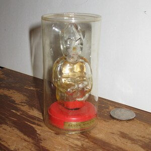 K&ouml;nnte beinhalten: Eine klare Glasfigur einer sitzenden Figur mit goldfarbenem Torso, eingeschlossen in einem transparenten Kunststoffzylinder. Die Figur sitzt auf einem roten Sockel mit dem Text "Devon Violets" auf einem goldenen Etikett. Eine M&uuml;nze liegt daneben.