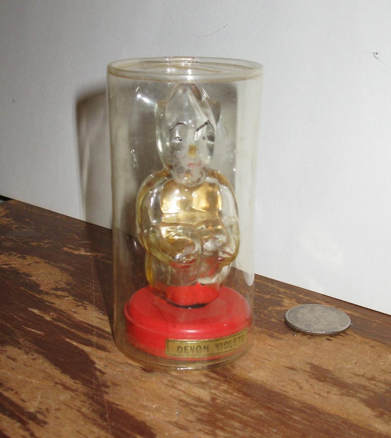 K&ouml;nnte beinhalten: Eine klare Glasfigur einer sitzenden Figur mit goldfarbenem Torso, eingeschlossen in einem transparenten Kunststoffzylinder. Die Figur sitzt auf einem roten Sockel mit dem Text "Devon Violets" auf einem goldenen Etikett. Eine M&uuml;nze liegt daneben.
