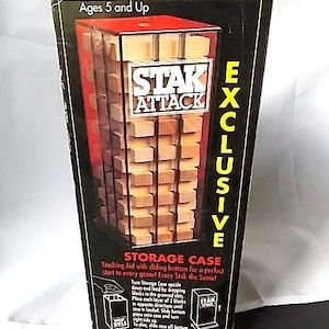 May include: A Stak Attack game box with a clear plastic window showcasing wooden blocks. The box is black with yellow and white text, including "EXCLUSIVE" and "STORAGE CASE." The game is for ages 5 and up.