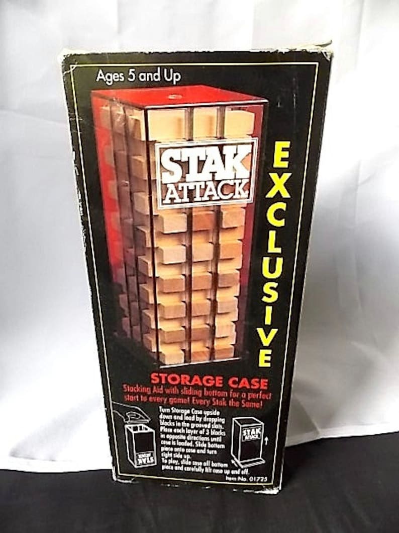 May include: A Stak Attack game box with a clear plastic window showcasing wooden blocks. The box is black with yellow and white text, including "EXCLUSIVE" and "STORAGE CASE." The game is for ages 5 and up.
