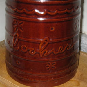 May include: A brown ceramic cookie jar with the word "cookies" embossed on the side. The jar has a cylindrical shape with decorative floral and wavy patterns. The jar is a deep reddish-brown color.