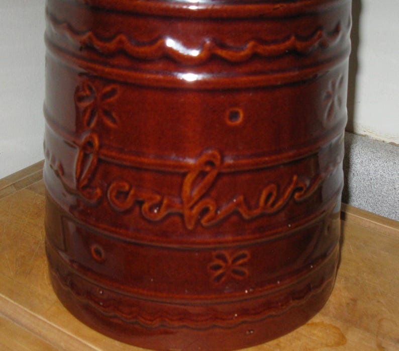 May include: A brown ceramic cookie jar with the word "cookies" embossed on the side. The jar has a cylindrical shape with decorative floral and wavy patterns. The jar is a deep reddish-brown color.