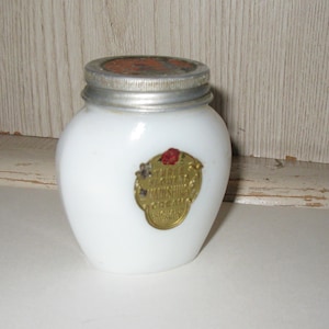 May include: A small, white, vintage jar with a silver lid and a gold label. The label has red and gold accents and text. The jar is likely for cosmetics or creams. The jar is sitting on a white surface.