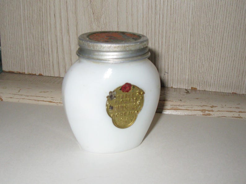 May include: A small, white, vintage jar with a silver lid and a gold label. The label has red and gold accents and text. The jar is likely for cosmetics or creams. The jar is sitting on a white surface.