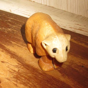 Peut inclure: Petite figurine d'ours en bois sculpt&eacute;, corps brun clair et t&ecirc;te cr&egrave;me. L'ours a des yeux noirs et est en position debout. La figurine est pos&eacute;e sur une surface en bois.