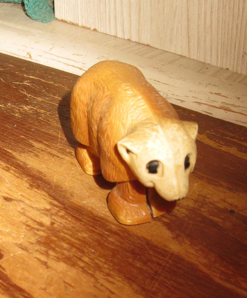 Peut inclure: Petite figurine d'ours en bois sculpt&eacute;, corps brun clair et t&ecirc;te cr&egrave;me. L'ours a des yeux noirs et est en position debout. La figurine est pos&eacute;e sur une surface en bois.