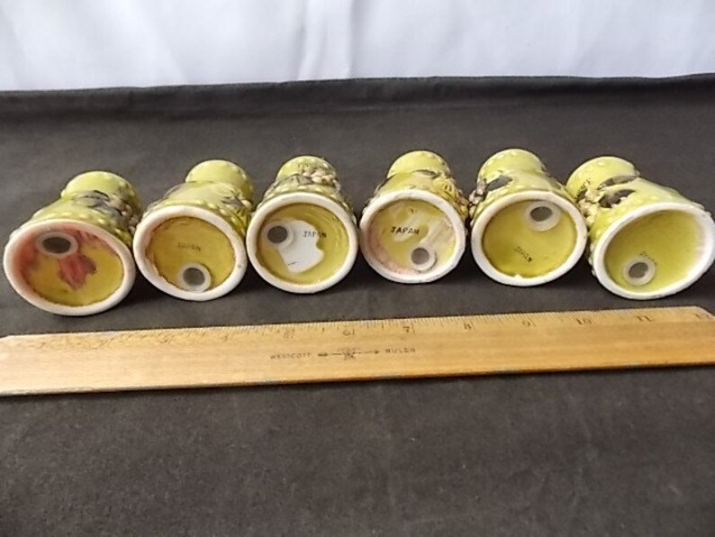 May include: Six vintage ceramic salt and pepper shakers. The shakers are yellow with black and brown floral designs. The bottoms are white with a single hole for dispensing. A wooden ruler is in the foreground.