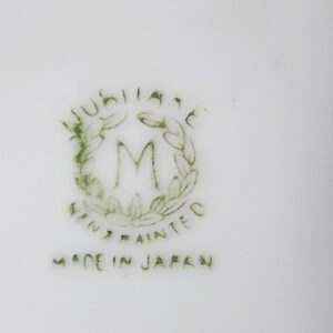 May include: A close-up of a white ceramic surface with a green and white emblem. The emblem features the word "Jubilee" above a stylized "M" within a wreath, with "Hand Painted" and "Made in Japan" below.
