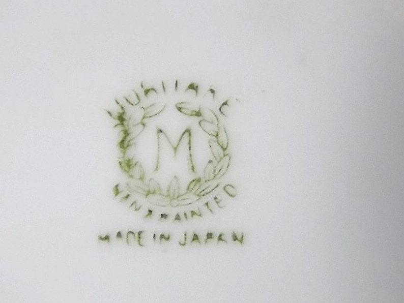 May include: A close-up of a white ceramic surface with a green and white emblem. The emblem features the word "Jubilee" above a stylized "M" within a wreath, with "Hand Painted" and "Made in Japan" below.