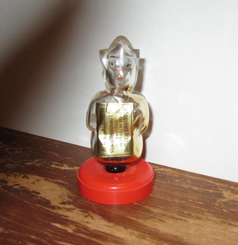 K&ouml;nnte beinhalten: Eine Vintage-Parf&uuml;mflasche in Form einer Figur, mit einem transparenten Glask&ouml;rper und einem roten Sockel. Die Flasche hat ein Etikett mit Text. Die Figur hat einen Hut und h&auml;lt die Arme eng am K&ouml;rper. Die Flasche steht auf einer Holzoberfl&auml;che.