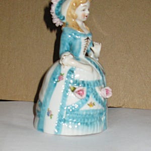 May include: A porcelain figurine of a person wearing a blue and white dress with a matching bonnet. The dress features blue vertical stripes, small floral patterns, and three-dimensional pink rose accents. The figure has blonde hair and a bodice.