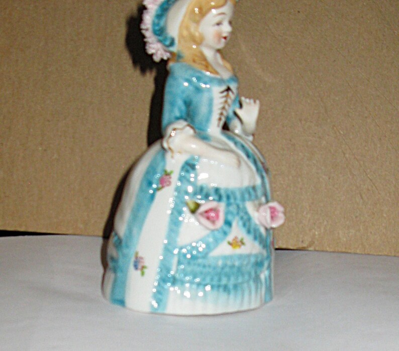 May include: A porcelain figurine of a person wearing a blue and white dress with a matching bonnet. The dress features blue vertical stripes, small floral patterns, and three-dimensional pink rose accents. The figure has blonde hair and a bodice.