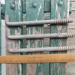 May include: Three gray metal L-shaped lag bolts with threaded ends. A wooden ruler is positioned horizontally beneath the bolts, showing that each bolt measures approximately 10 inches in length from the bent end to the tip of the screw thread.