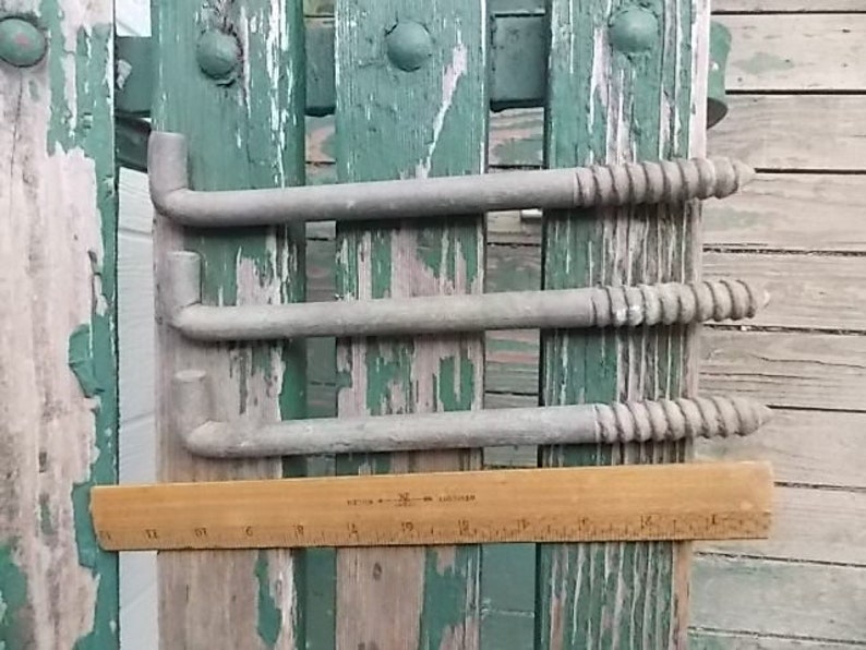 May include: Three gray metal L-shaped lag bolts with threaded ends. A wooden ruler is positioned horizontally beneath the bolts, showing that each bolt measures approximately 10 inches in length from the bent end to the tip of the screw thread.