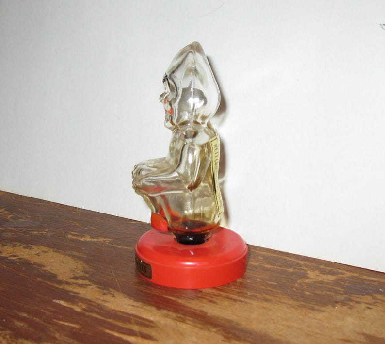 K&ouml;nnte beinhalten: Eine Vintage-Glasflasche in Form eines sitzenden Zwergs mit rotem Sockel. Die klare Glasfigur hat eine spitze M&uuml;tze und ein l&auml;chelndes Gesicht. Auf der R&uuml;ckseite der Flasche ist ein kleines Etikett angebracht. Der Sockel ist leuchtend rot.