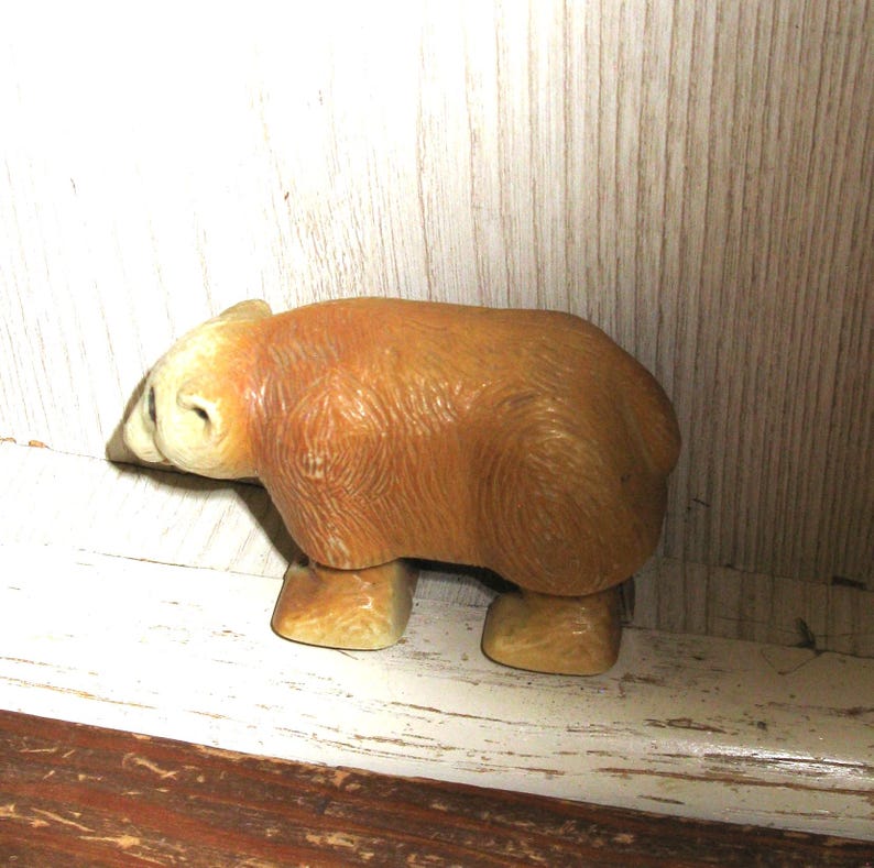 Peut inclure: Petite figurine d'ours brun avec une t&ecirc;te et des pattes de couleur claire. L'ours a une surface textur&eacute;e, lui donnant un aspect r&eacute;aliste. La figurine est expos&eacute;e sur une surface en bois blanc vieilli.