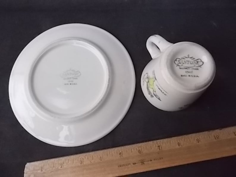 May include: White ceramic teacup and saucer set. The teacup has a handle and a decorative bird design. The saucer and the bottom of the teacup have the text "SANTONE" and "MADE IN U.S.A."